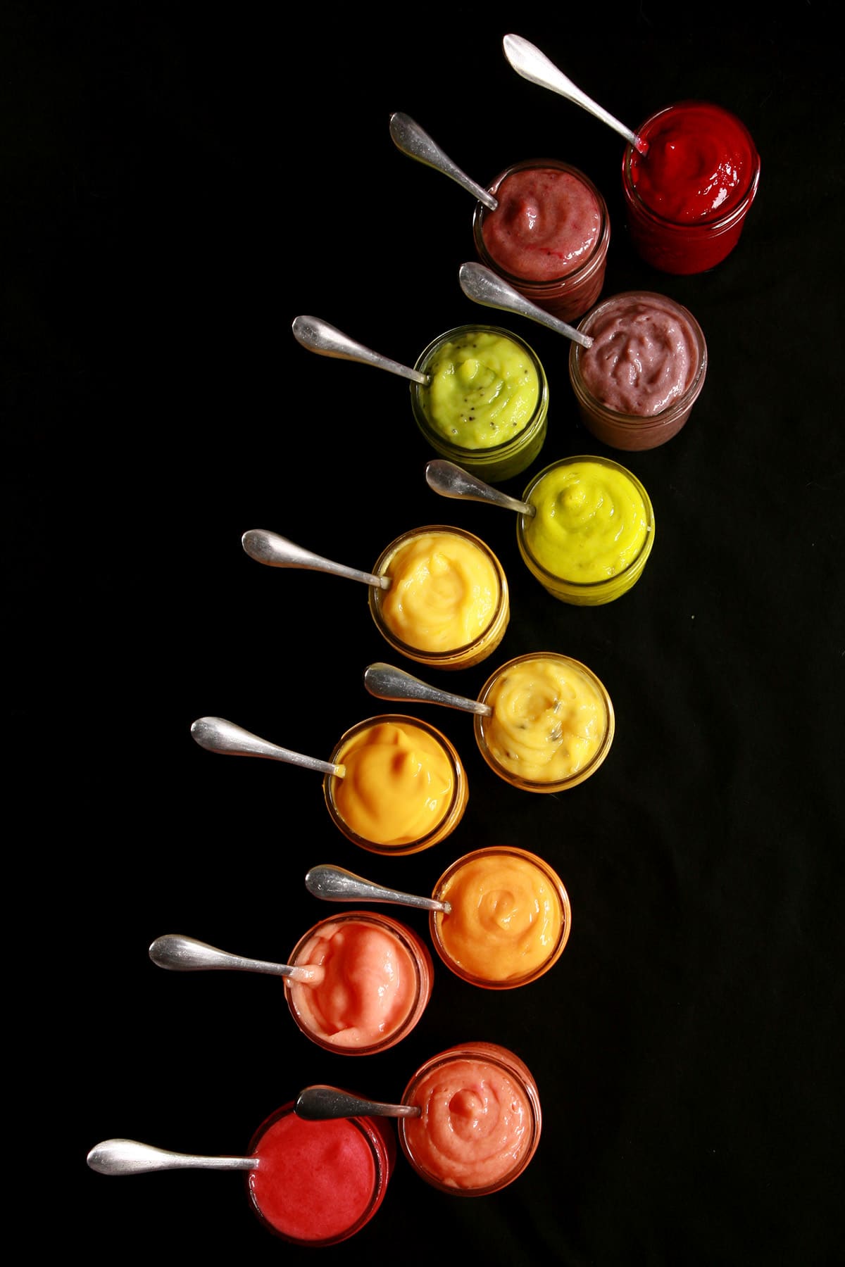 A line of open jars of fruit curd. There is a spoon in each, and they are arranged in rainbow order.