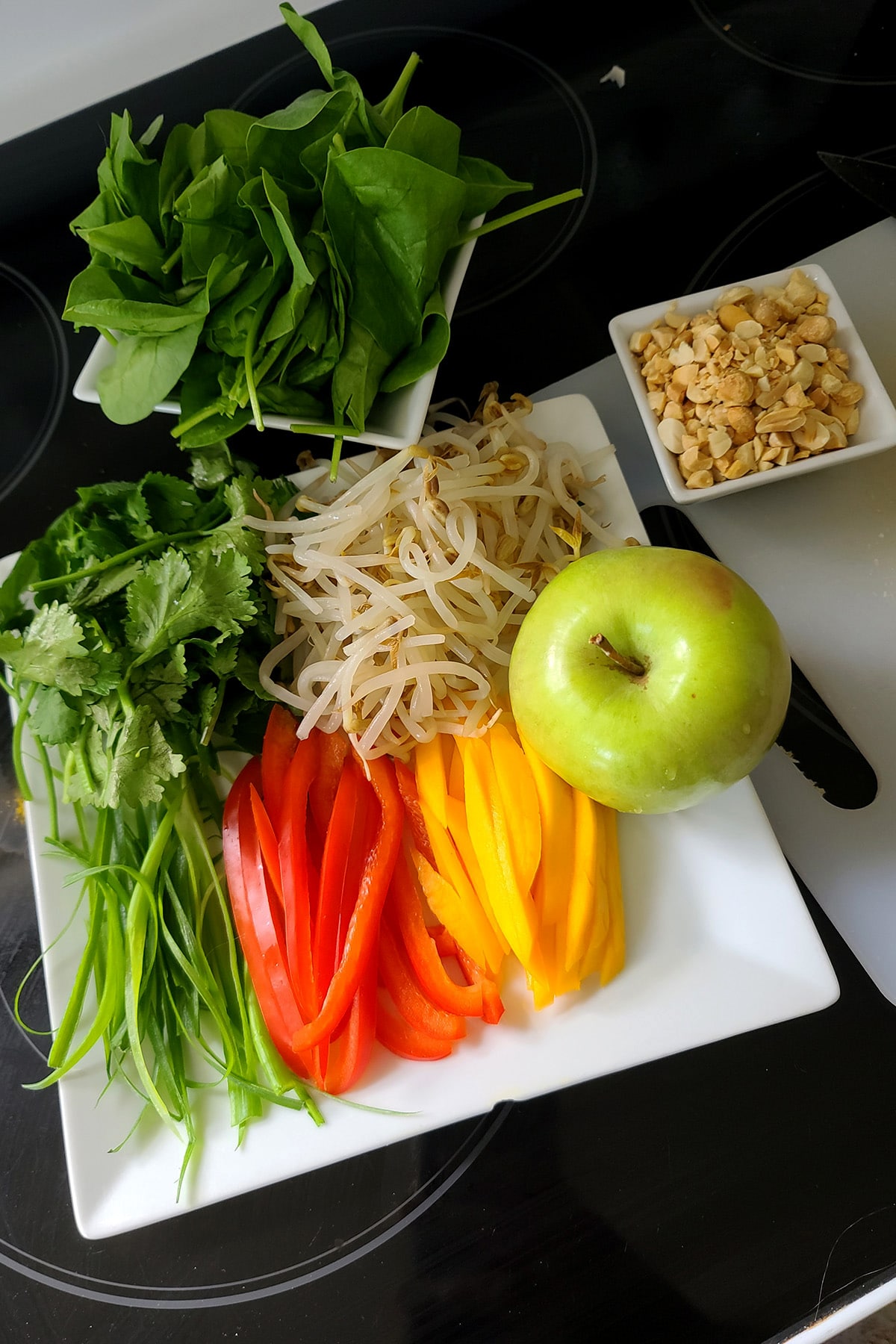 A plate with a green apple and slices of mango, red pepper, and green onions, along with bean sprouts, mint, peanuts, and spinach.