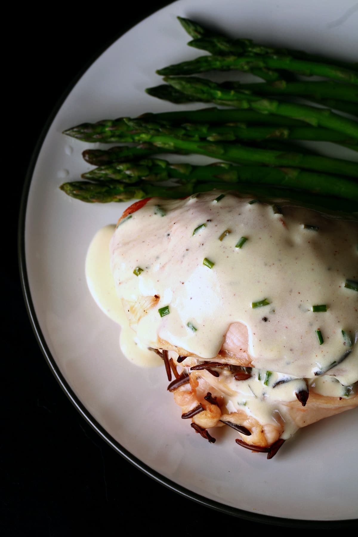 A wild rice stuffed chicken breast on a plate. It is covered in a Dijon chive cream sauce, and there are asparagus spears beside it.