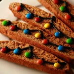 A white plate with several pieces of decorated gingerbread biscotti. Black frosting and mini M&Ms form a swag of christmas lights across each cookie.