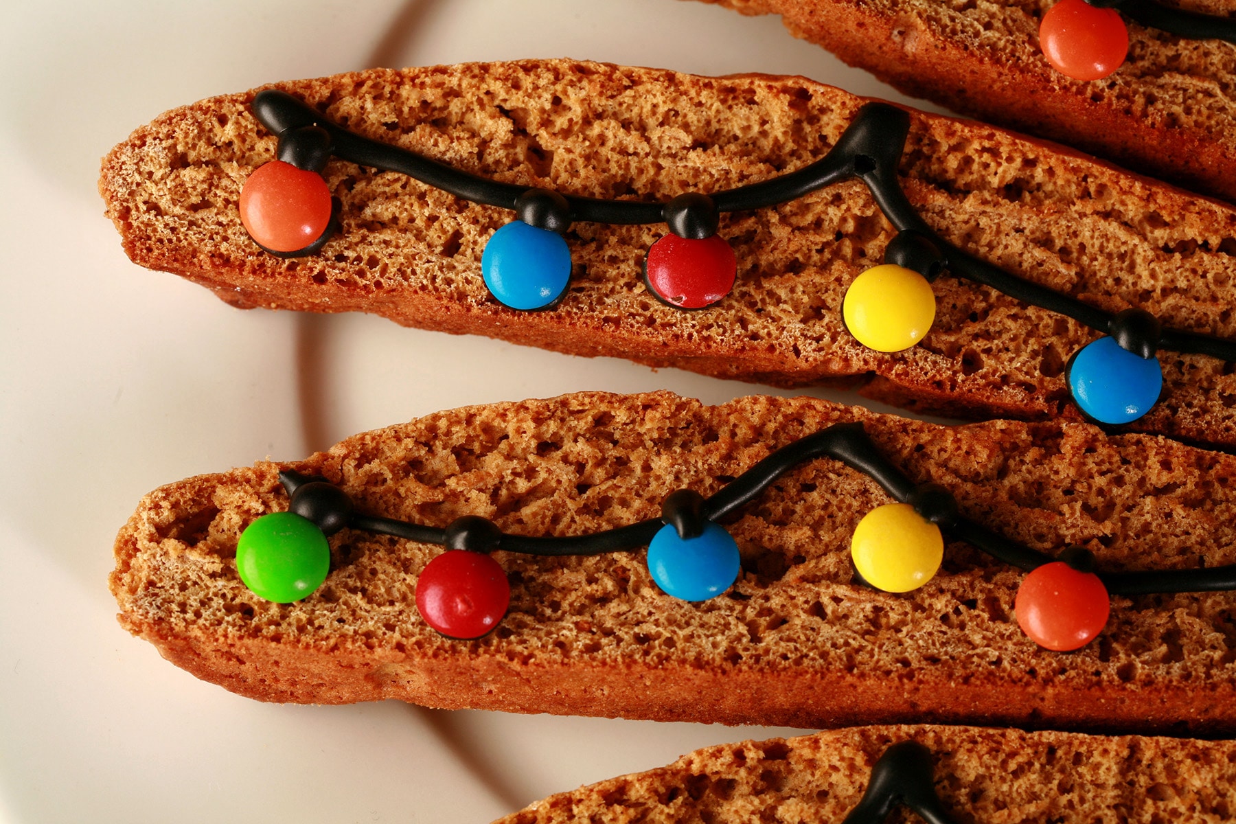 A white plate with several pieces of decorated gingerbread biscotti. Black frosting and mini M and Ms form a swag of christmas lights across each cookie.