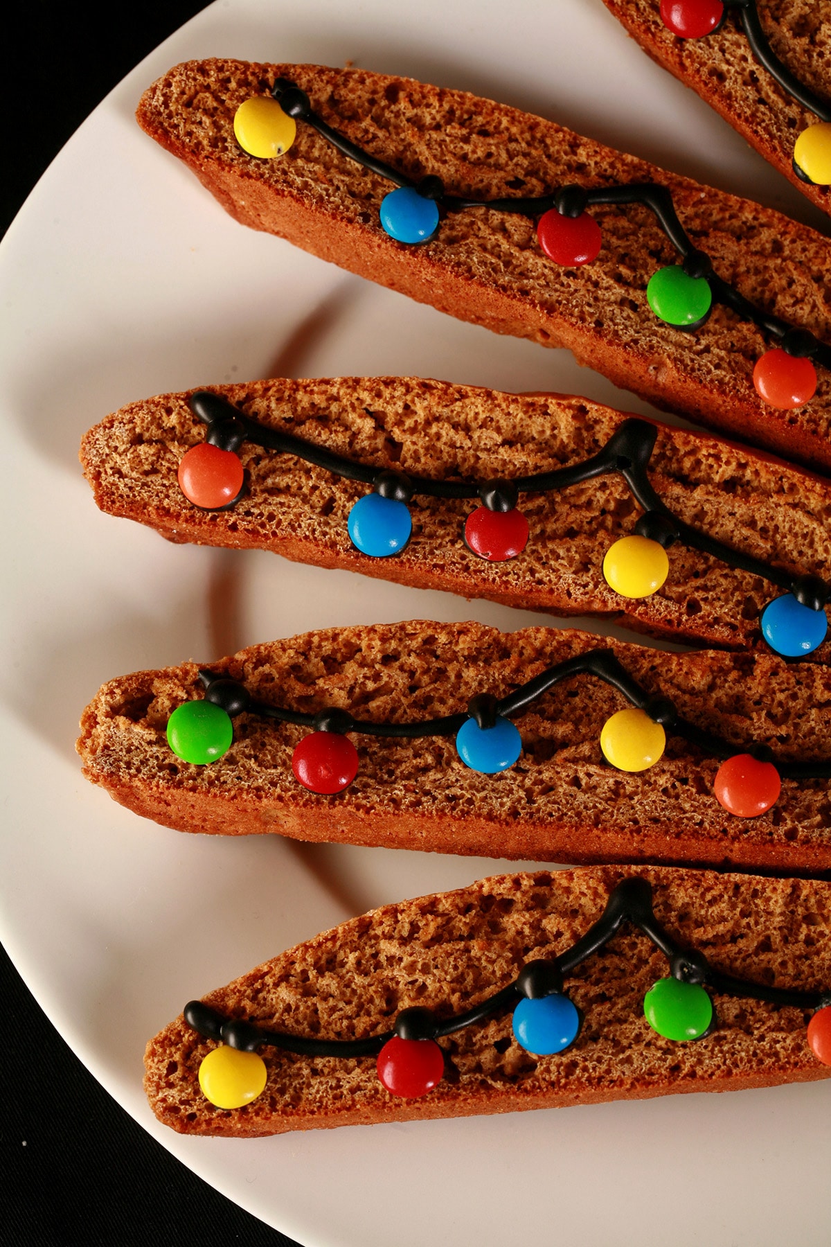 A white plate with several pieces of decorated gingerbread biscotti. Black frosting and mini M&Ms form a swag of christmas lights across each cookie.