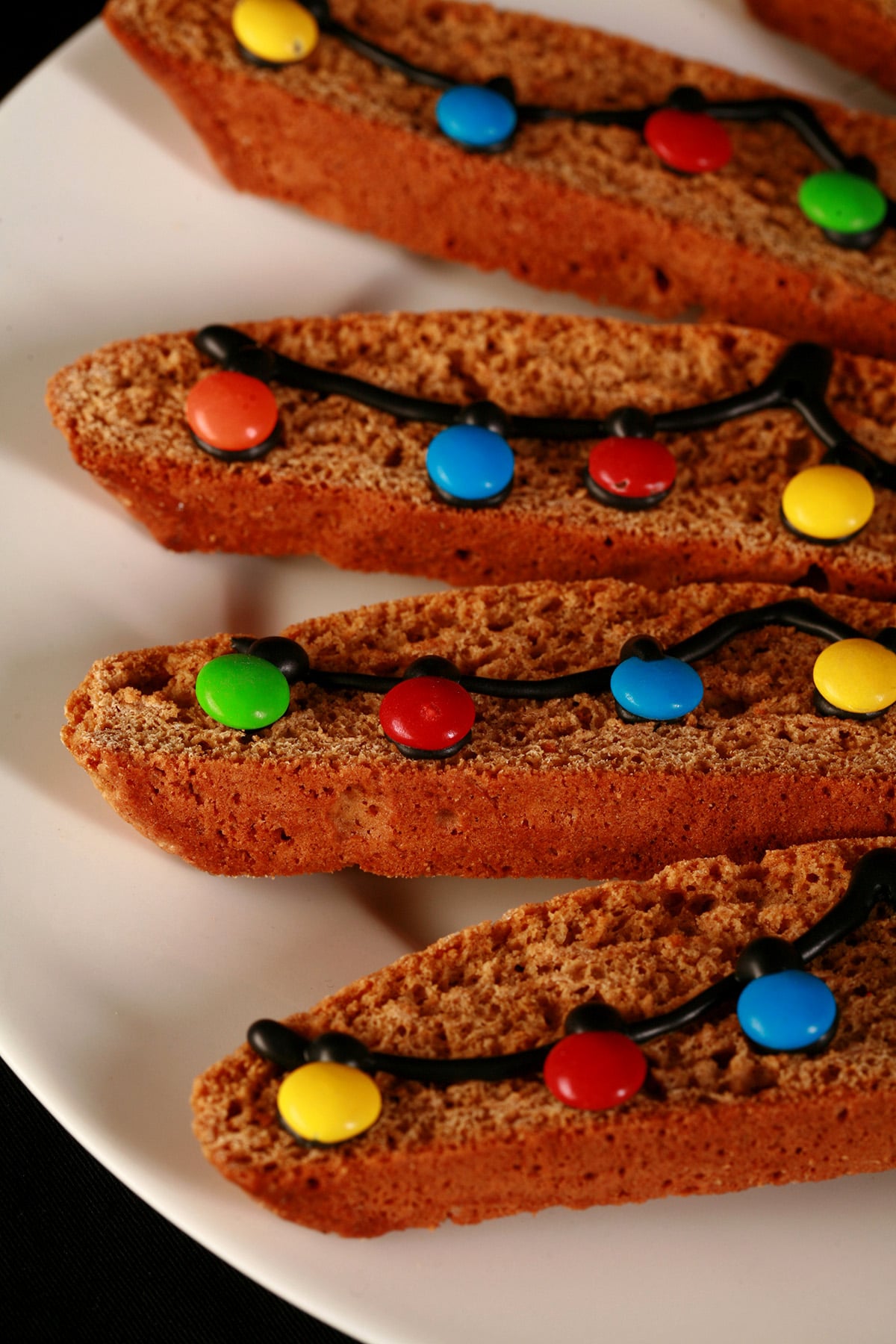 A white plate with several pieces of decorated gingerbread biscotti. Black frosting and mini M and Ms form a swag of christmas lights across each cookie.