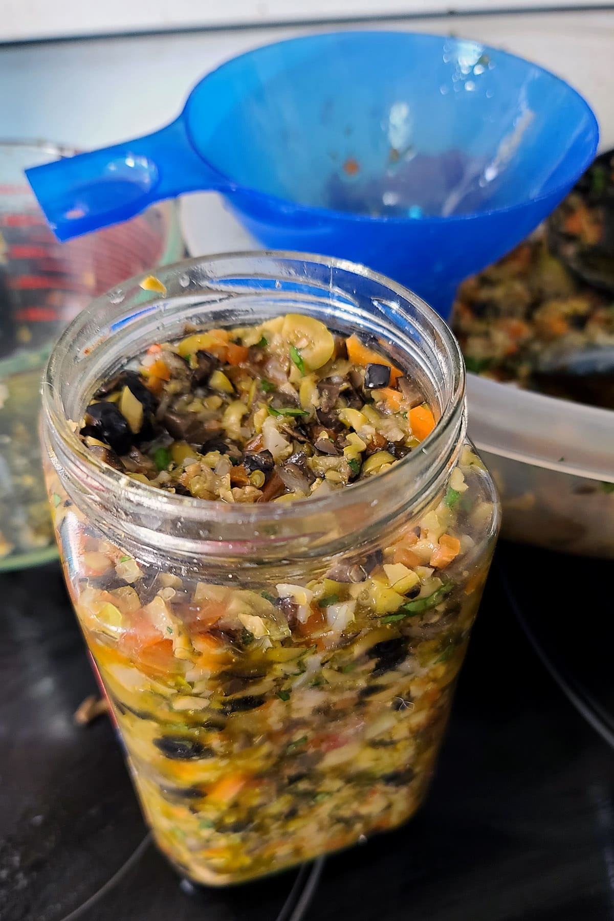 A large glass jar filled with olive salad, a funnel in the background.