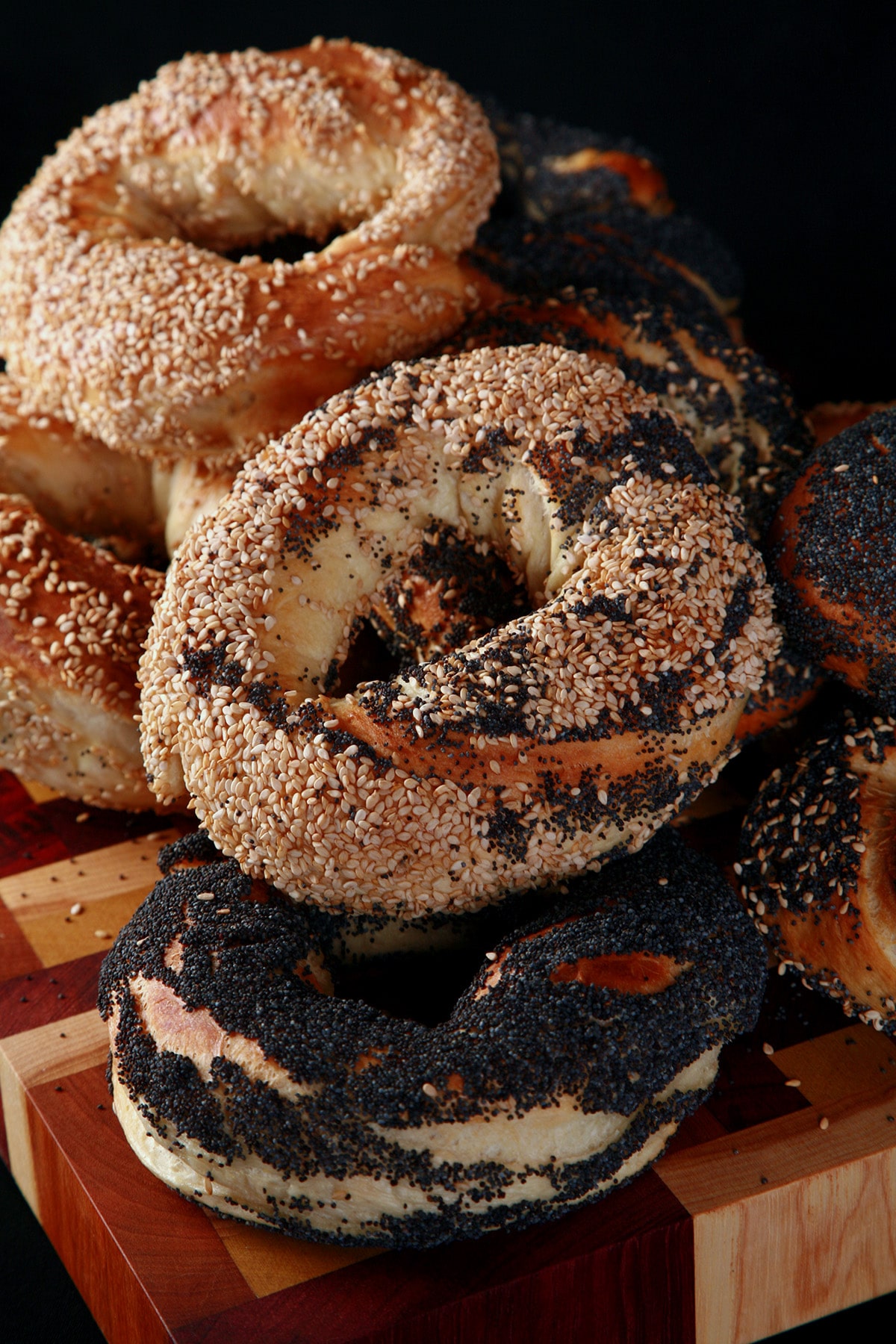 An assortment of Montreal Bagels piled on a cutting board. They are coated in sesame seeds and poppy seeds.