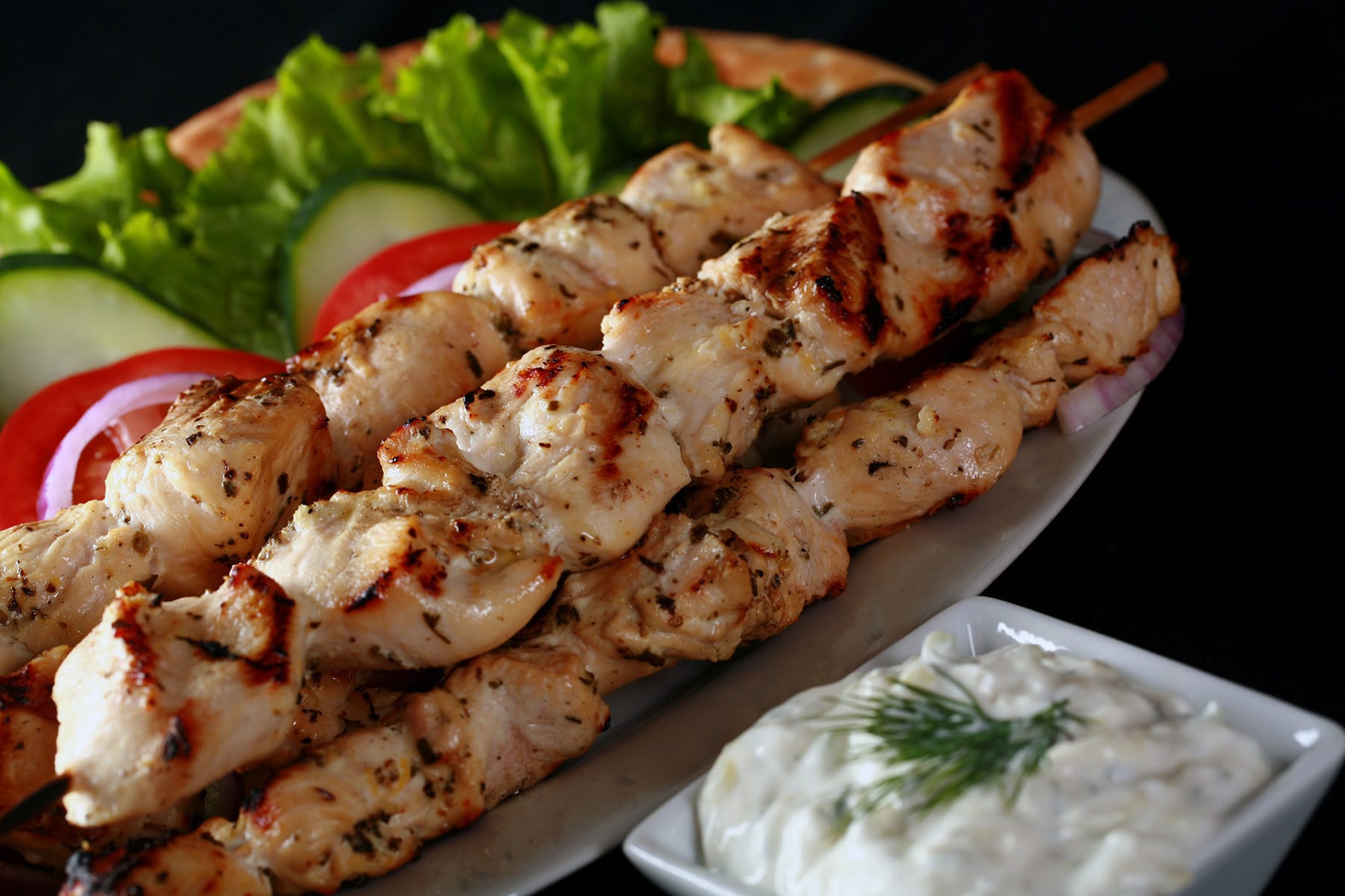 Several grilled chicken soulaki skewers on a platter with red onions, tomatoes, and cucumber slices.