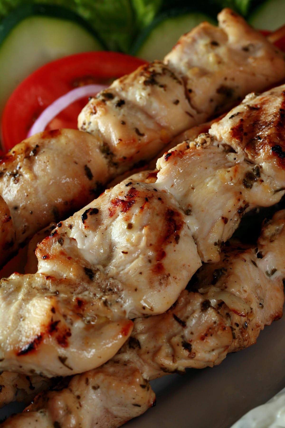 Several grilled chicken soulaki skewers on a platter with red onions, tomatoes, and cucumber slices.