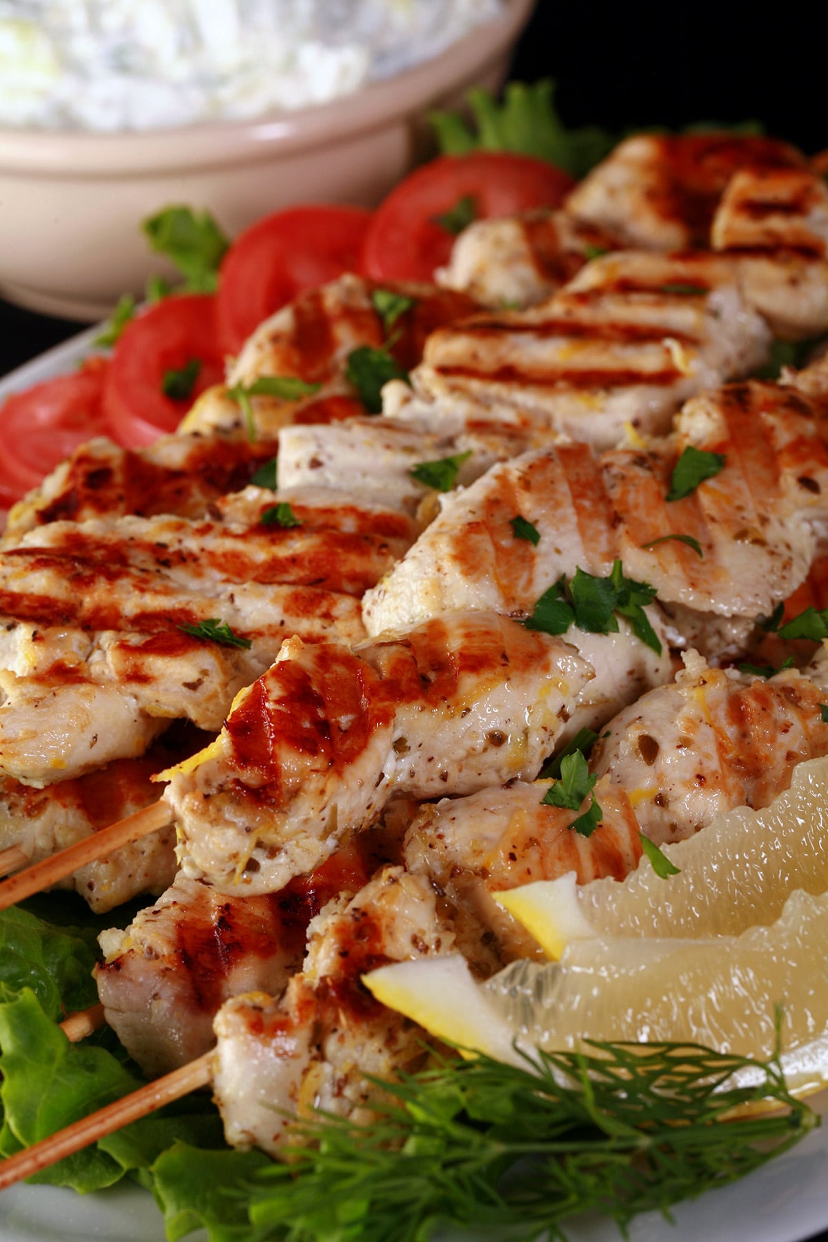 Several skewers of Green chicken souvlaki on a bed of lettuce, with tomatoes and lemon slices.