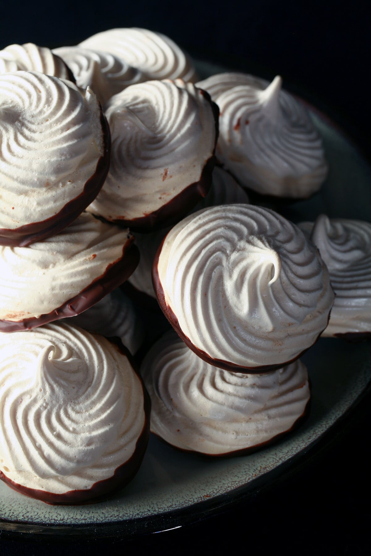A plate piled high with swirl shaped malted milk meringue cookies. The bottom of each one is coated in chocolate.