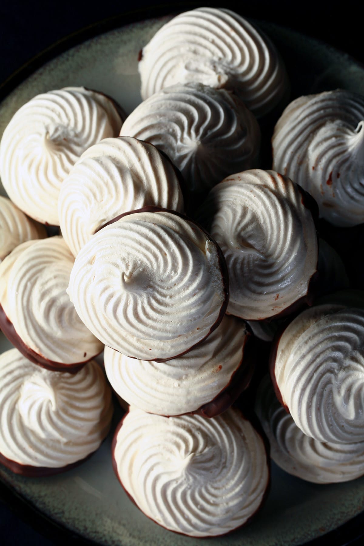 A plate piled high with swirl shaped malted milk meringue cookies. The bottom of each one is coated in chocolate.