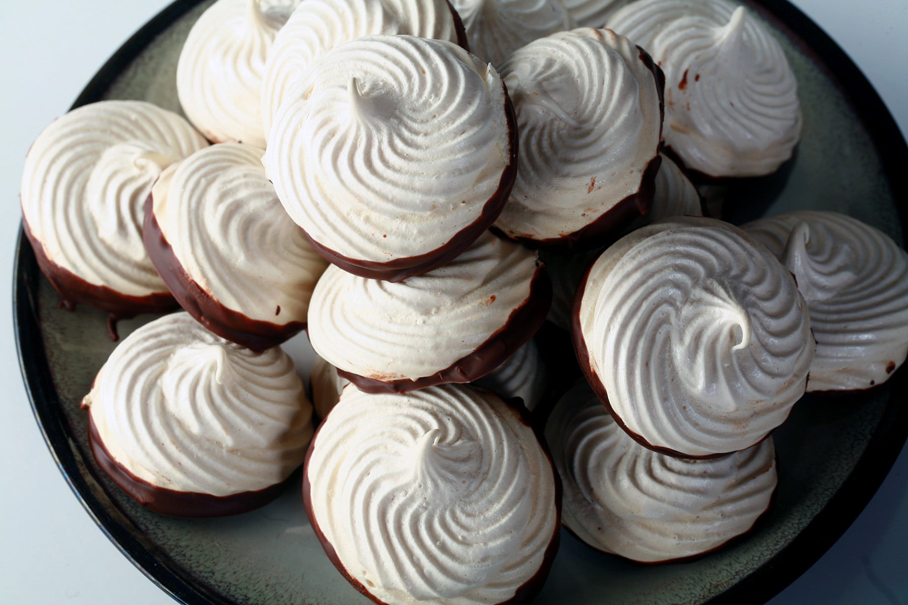 A plate piled high with swirl shaped malted milk meringue cookies. The bottom of each one is coated in chocolate.