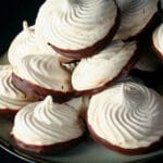 A plate piled high with swirl shaped malted milk meringue cookies. The bottom of each one is coated in chocolate.