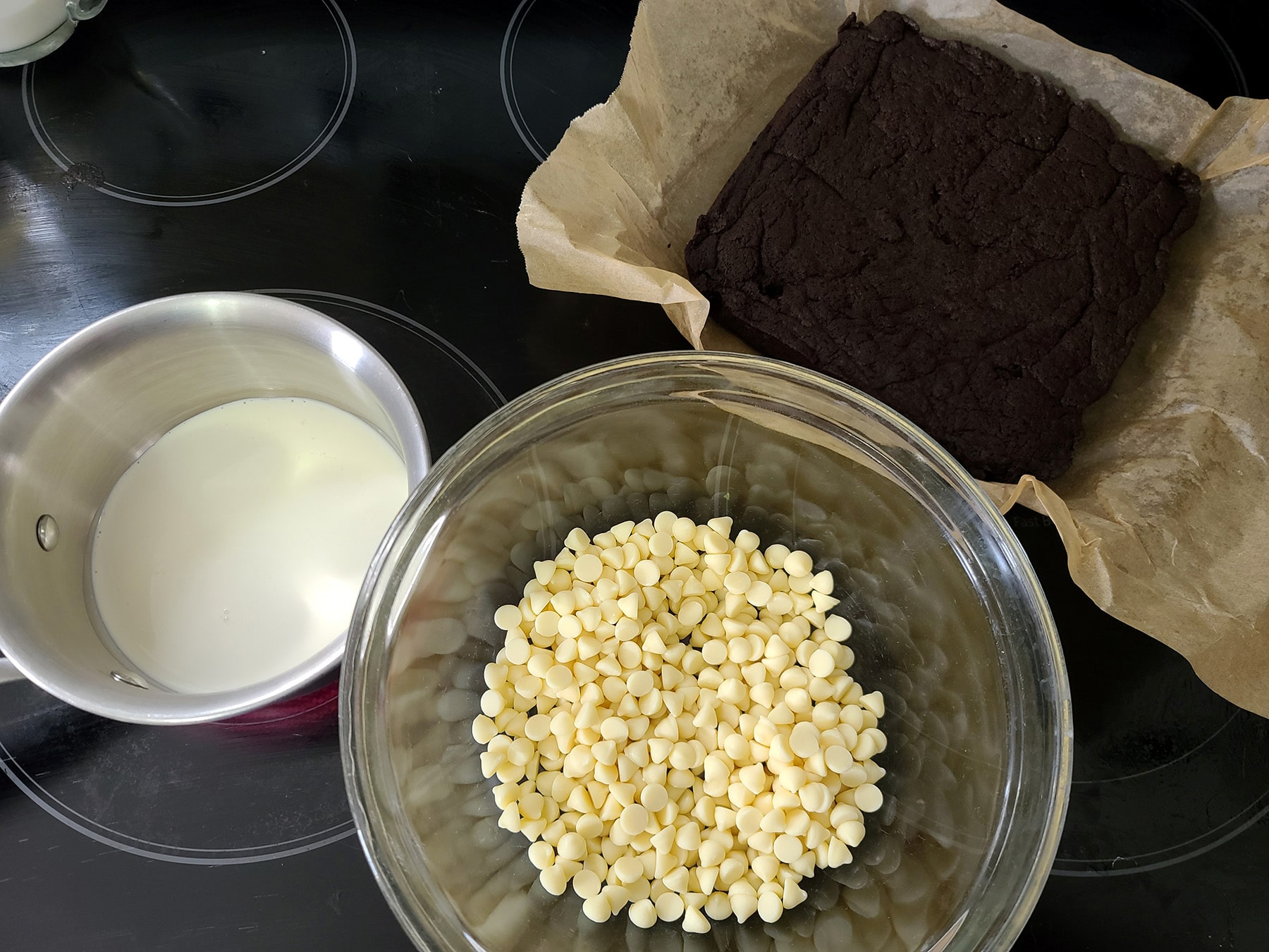 A bowl of white chocolate chips, a slab of brownie, and a small pot of heavy cream on a stovetop.