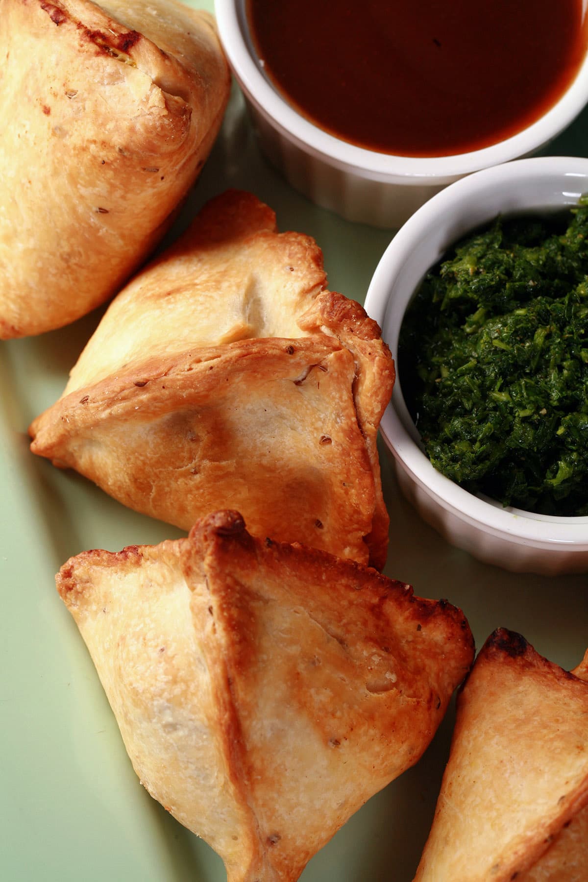 A plate of air fried samosas, with bowls of tamarind and mint chutneys beside them.