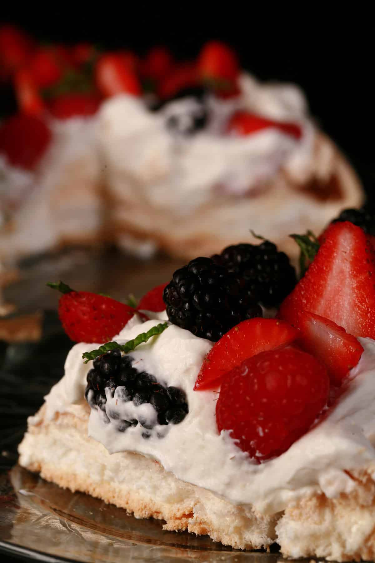 A slice of berry pavlova on a plate, with the remaining pavlova behind it.