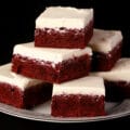 A plate stacked with red relvet brownies, each frosted with cream cheese icing.
