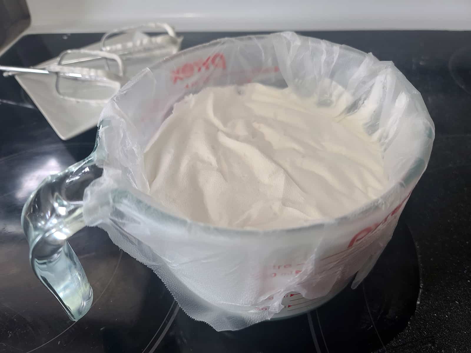 A large measuring cup of royal icing, covered with plastic wrap.