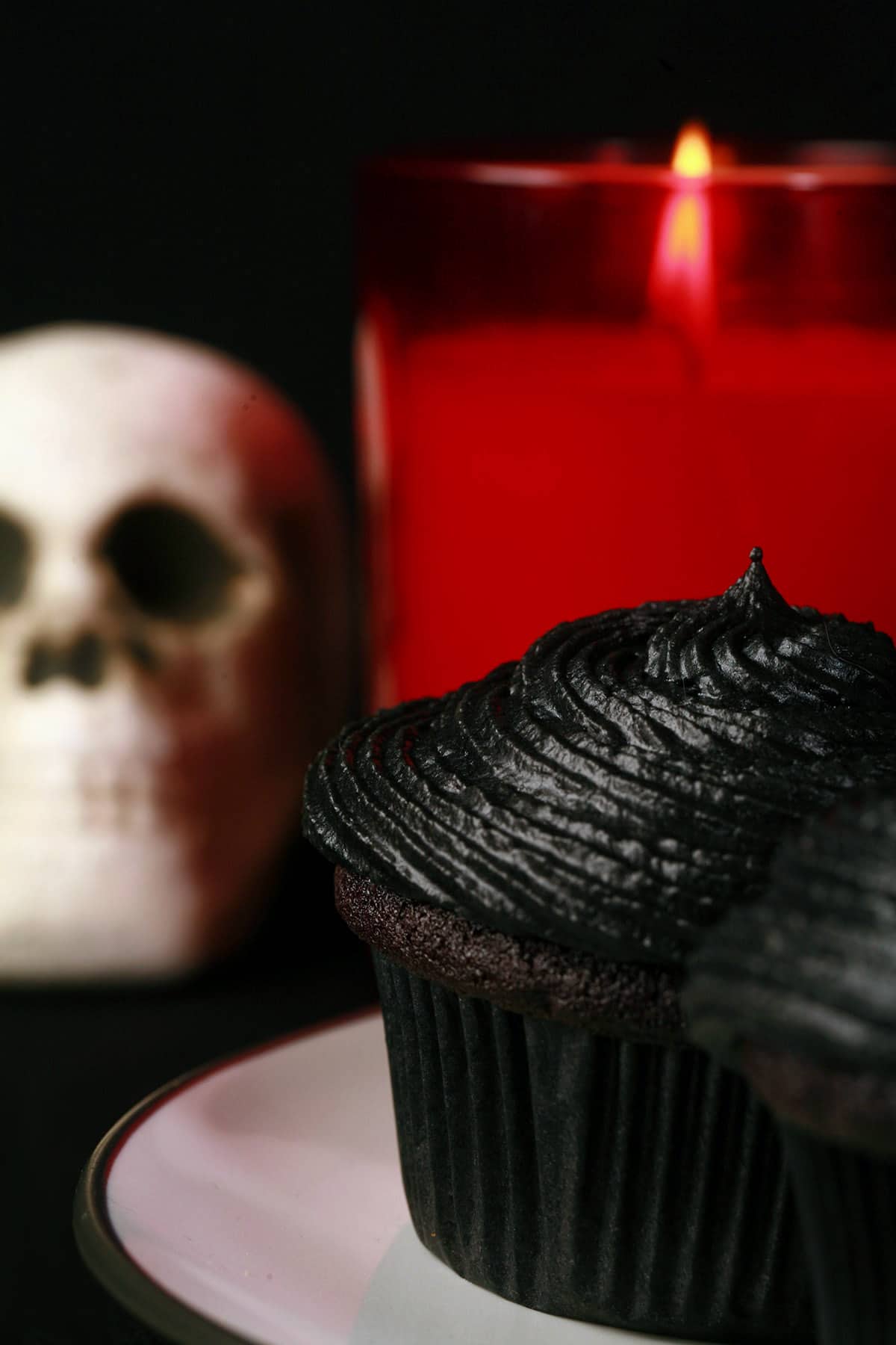A black velvet cupcake on a white plate, with a skull and red candle in the background.