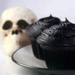 2 black velvet cupcakes on a white plate, with a skull in the background.