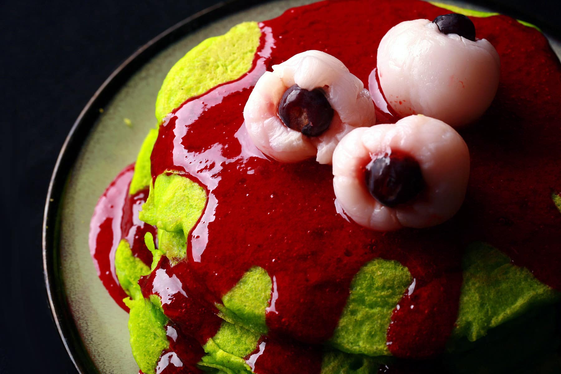 A stack of neon green Halloween pancakes with red sauce and lychee eyeballs on top.