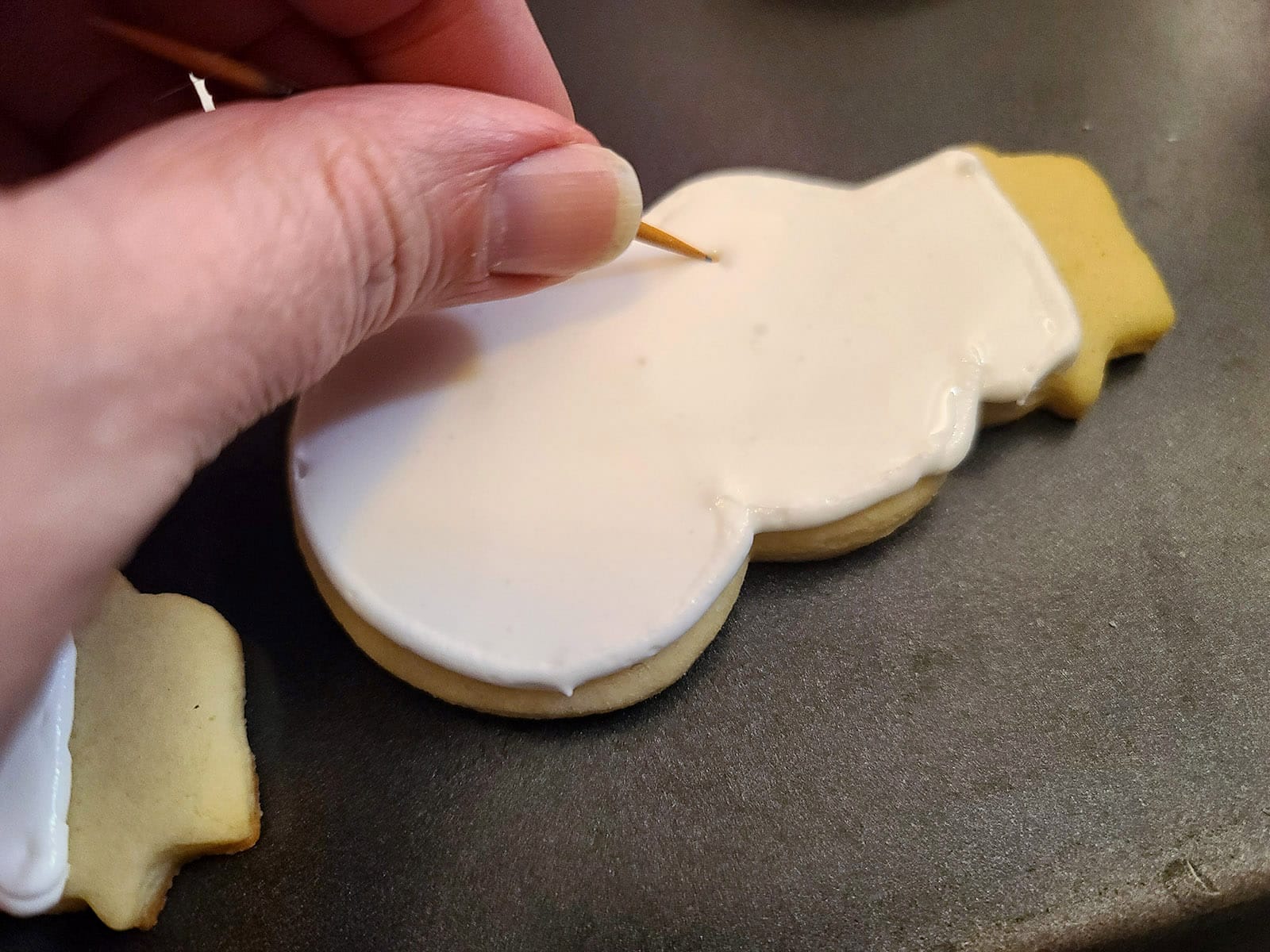 A hand uses a toothpick to pop bubbles in a snowman cookie frosted white.