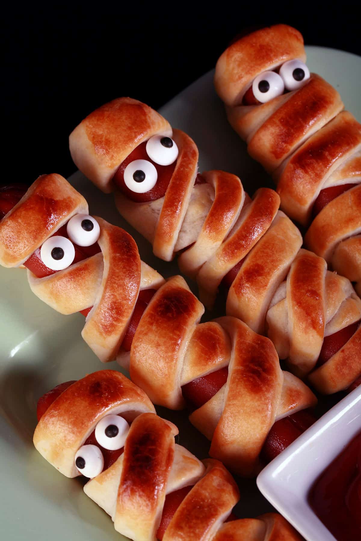 Several mummy dogs on a plate, each has 2 candy eyeballs.