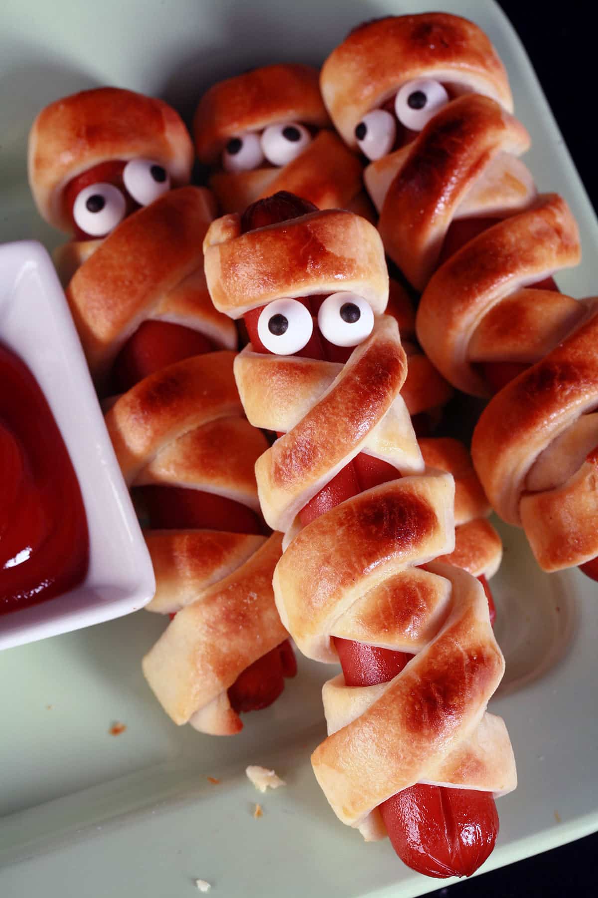 Several mummy dogs on a plate, each has 2 candy eyeballs.