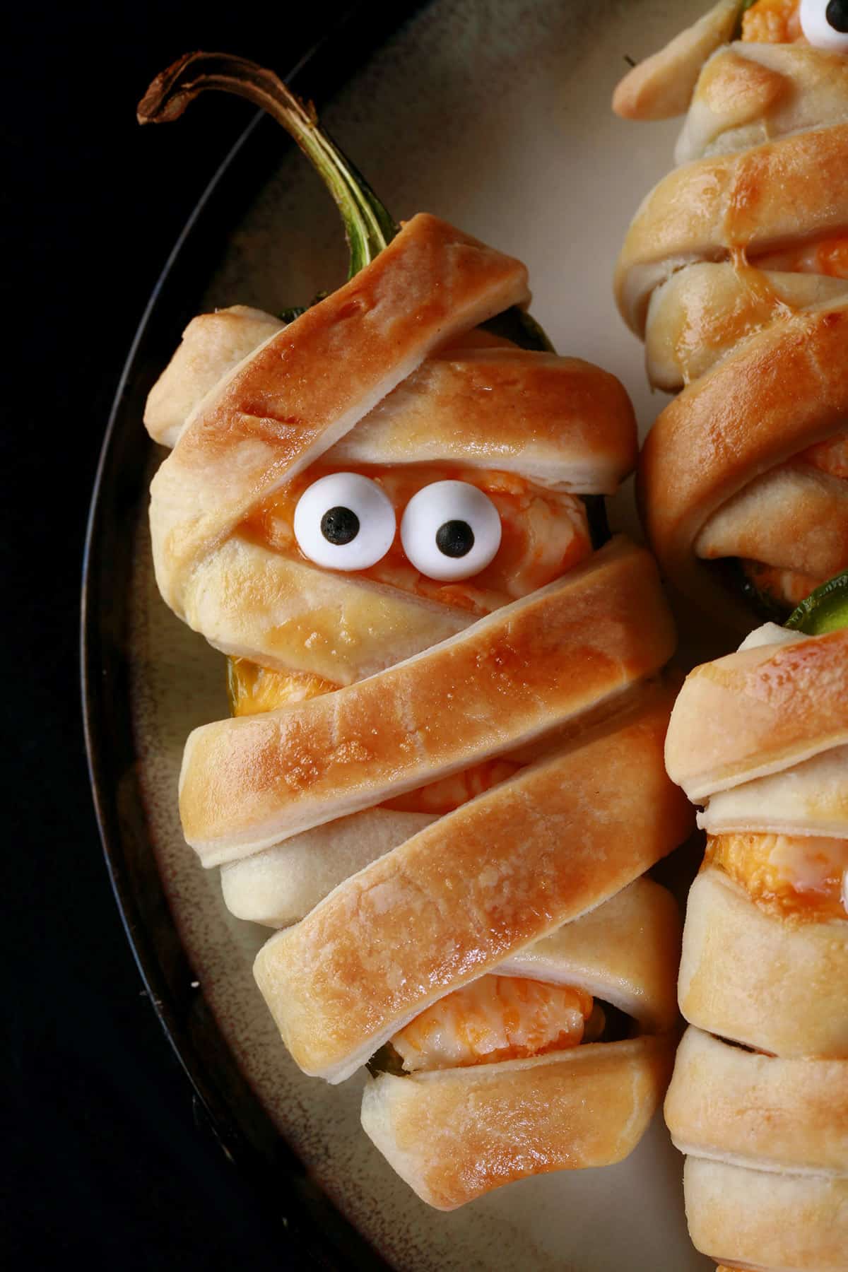 A plate of pastry wrapped Halloween mummy jalapeno poppers, each with 2 candy eyes.