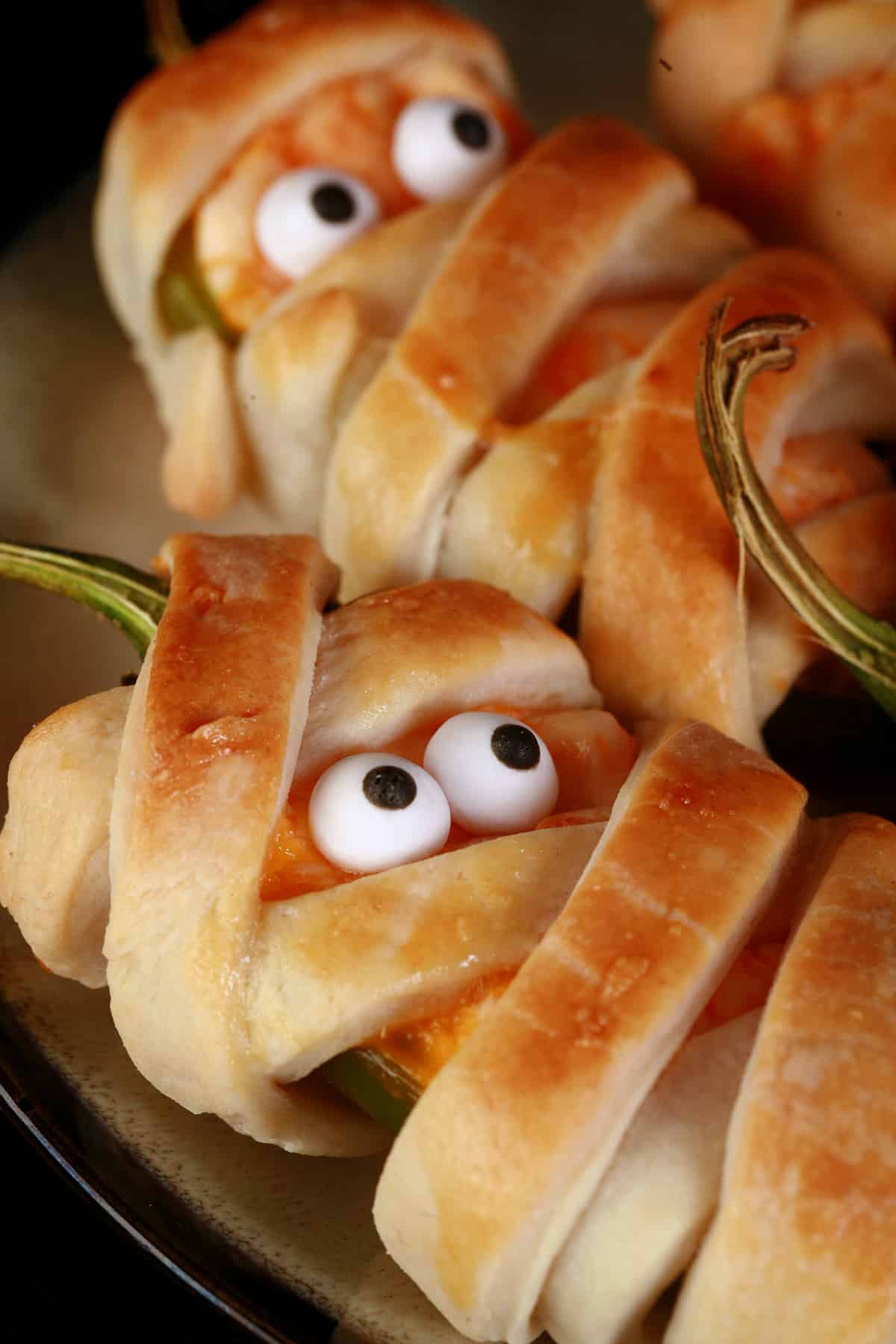 A plate of pastry wrapped Halloween mummy jalapeno poppers, each with 2 candy eyes.