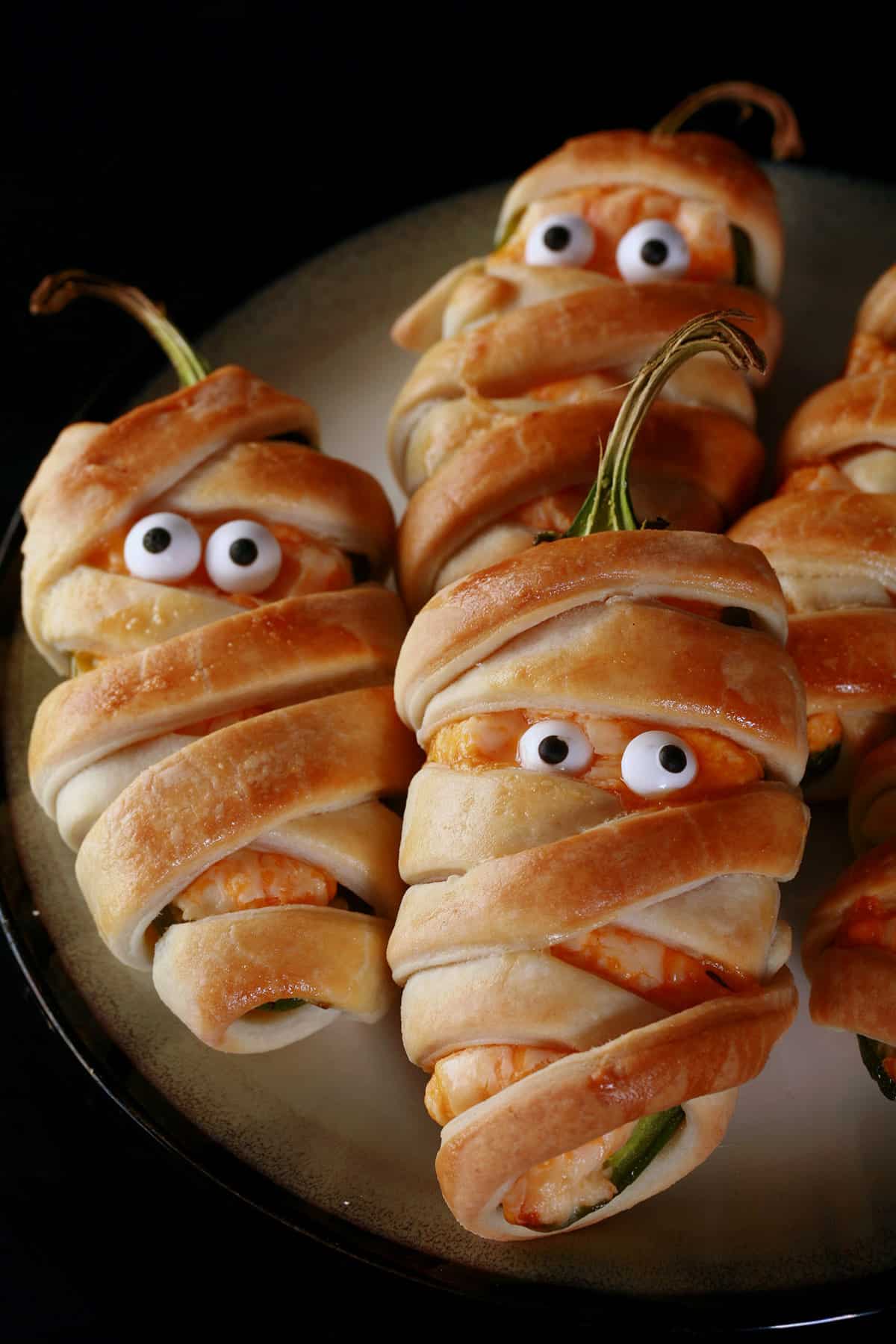 A plate of pastry wrapped Halloween mummy jalapeno poppers, each with 2 candy eyes.