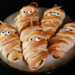 A plate of pastry wrapped Halloween mummy jalapeno poppers, each with 2 candy eyes.