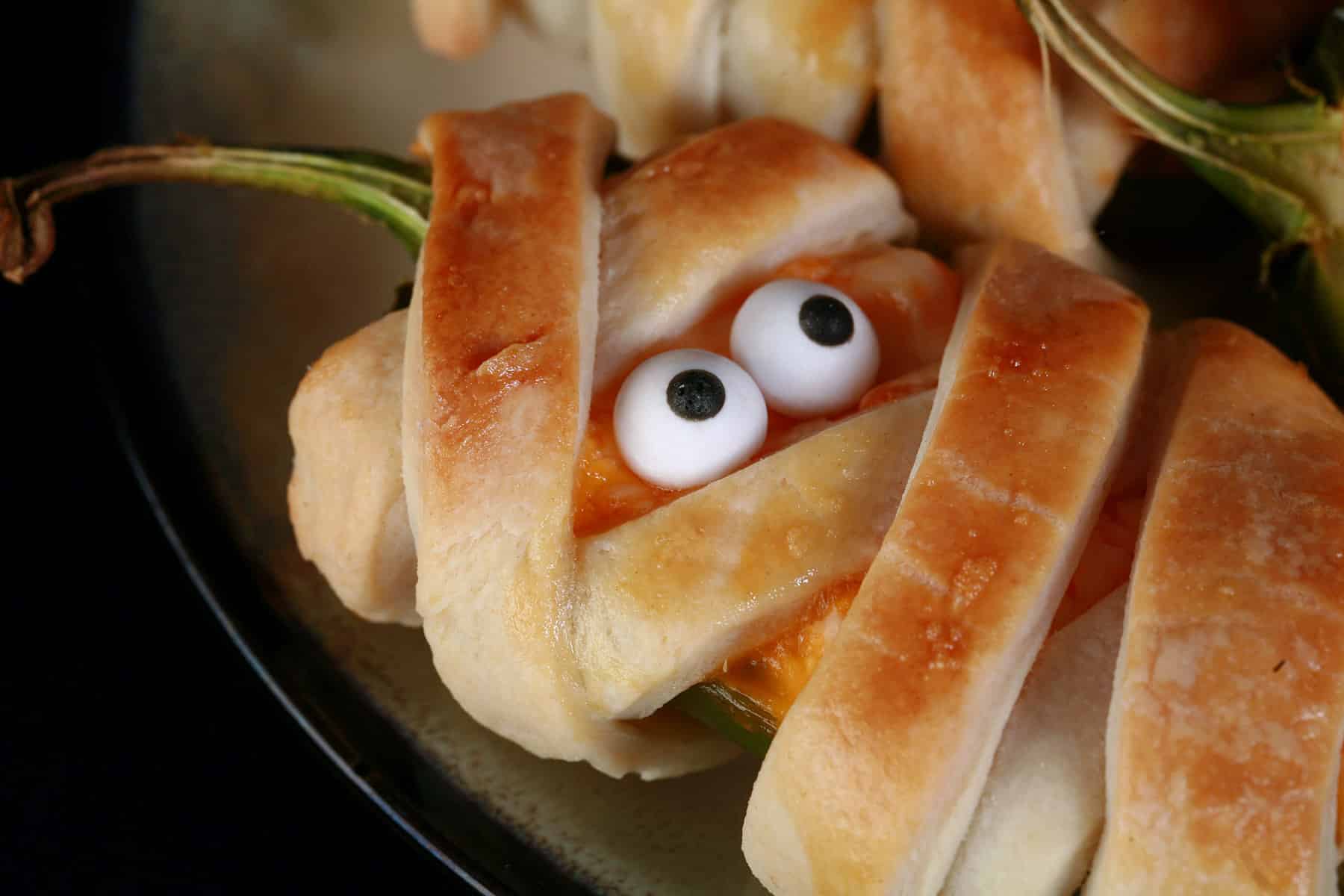 A plate of pastry wrapped Halloween mummy jalapeno poppers, each with 2 candy eyes.