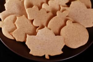 A plate of rolled cut-out pumpkin spice cookies in apple, pumpkin, and leaf shapes.