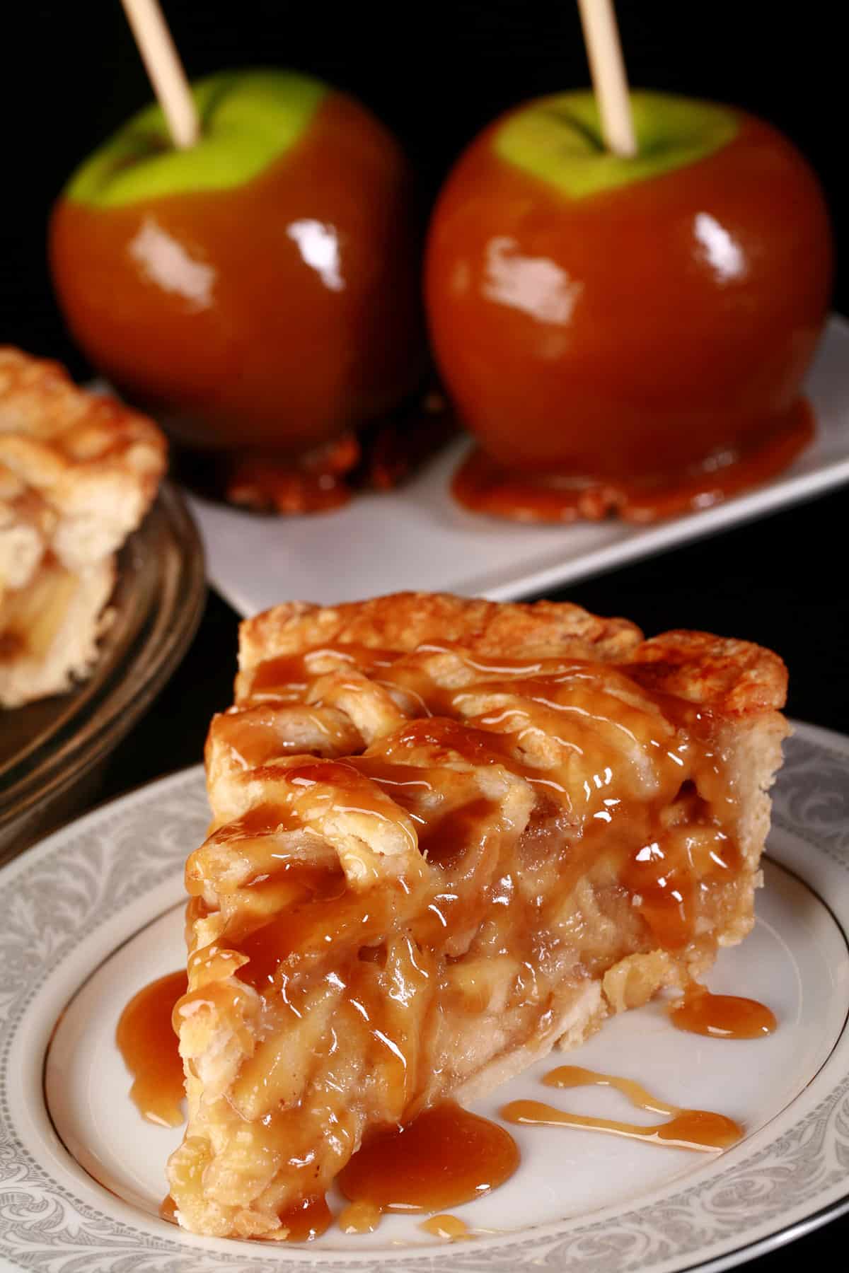 A slice of maple caramel apple pie on a plate in front of the rest of the pie and 2 caramel apples..