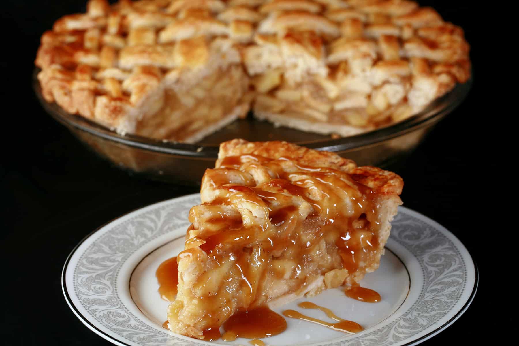 A slice of maple caramel apple pie on a plate in front of the rest of the pie.