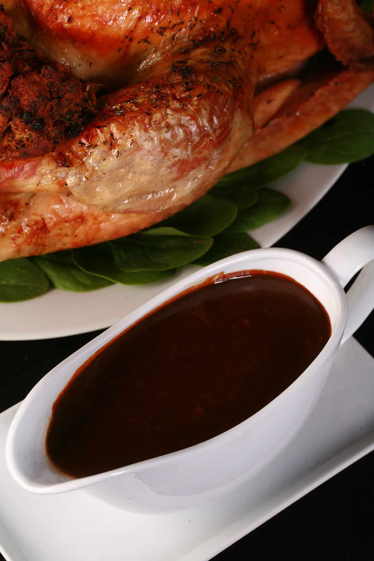 A gravy boat filled with very dark turkey gravy in front of a roasted stuffed turkey.