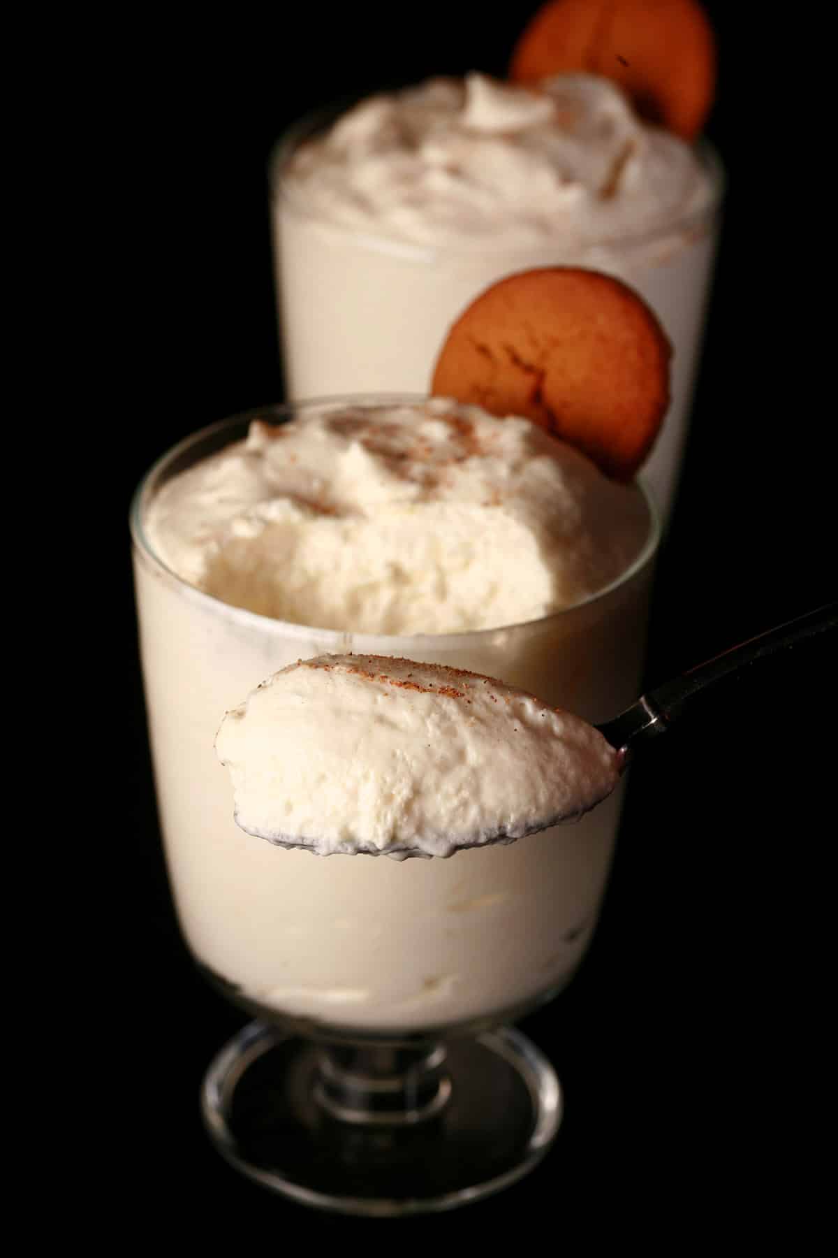 2 glasses of traditional style eggnog mousse, with nutmeg and gingersnap cookie garnish, with a spoon of mousse held in front.