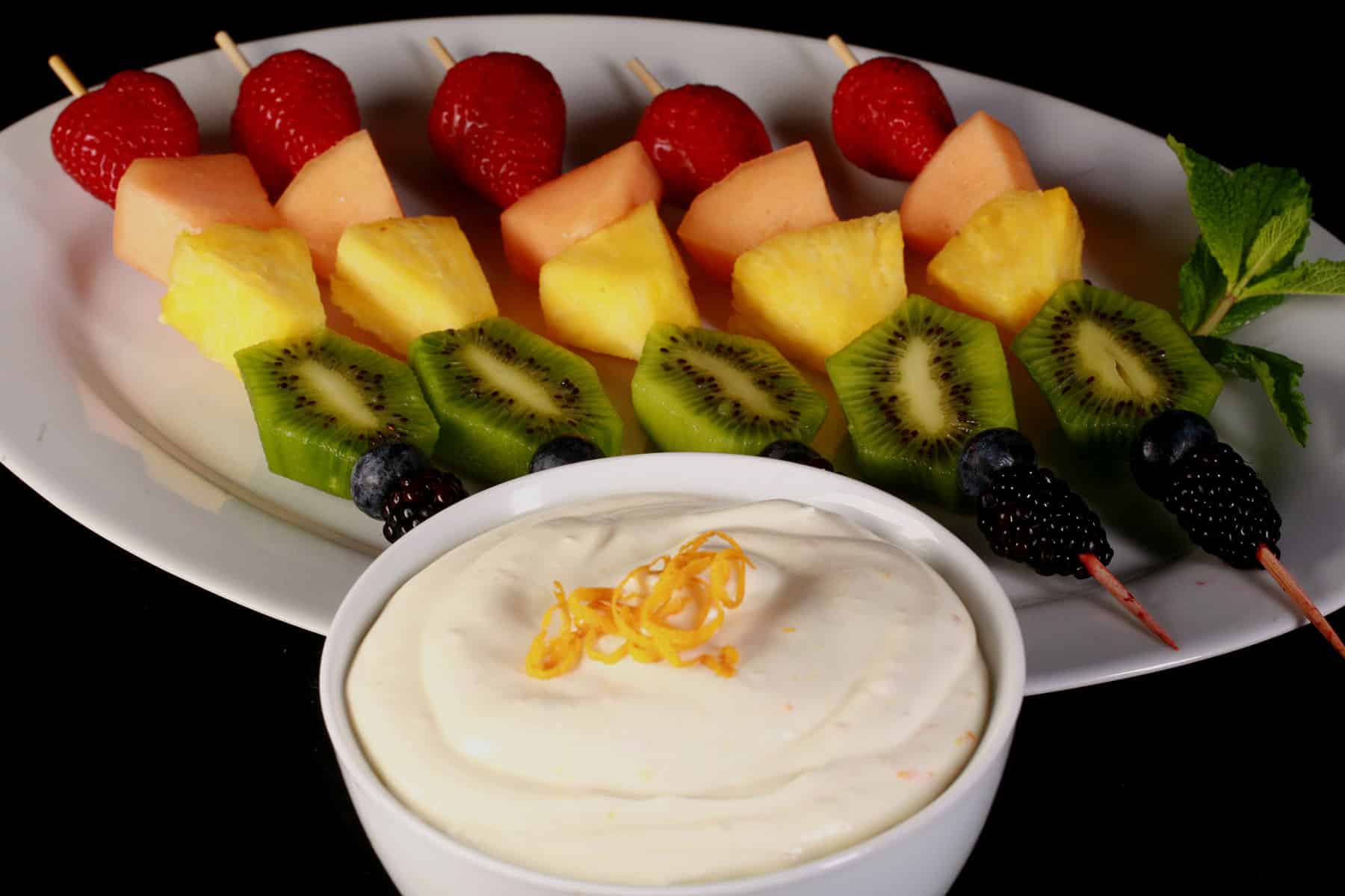 A platter of rainbow fruit skewers next to a bowl of mimosa cheesecake dip.