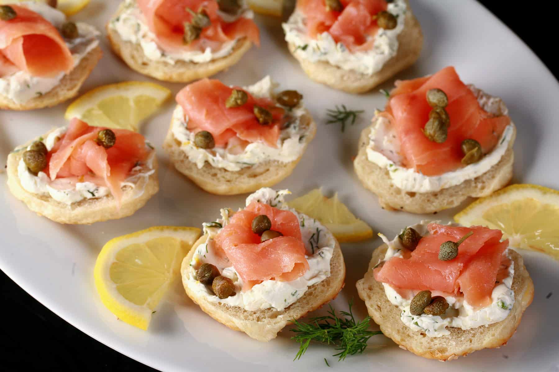 A platter of smoked salmon bagel bites garnished with capers and lemon slices.