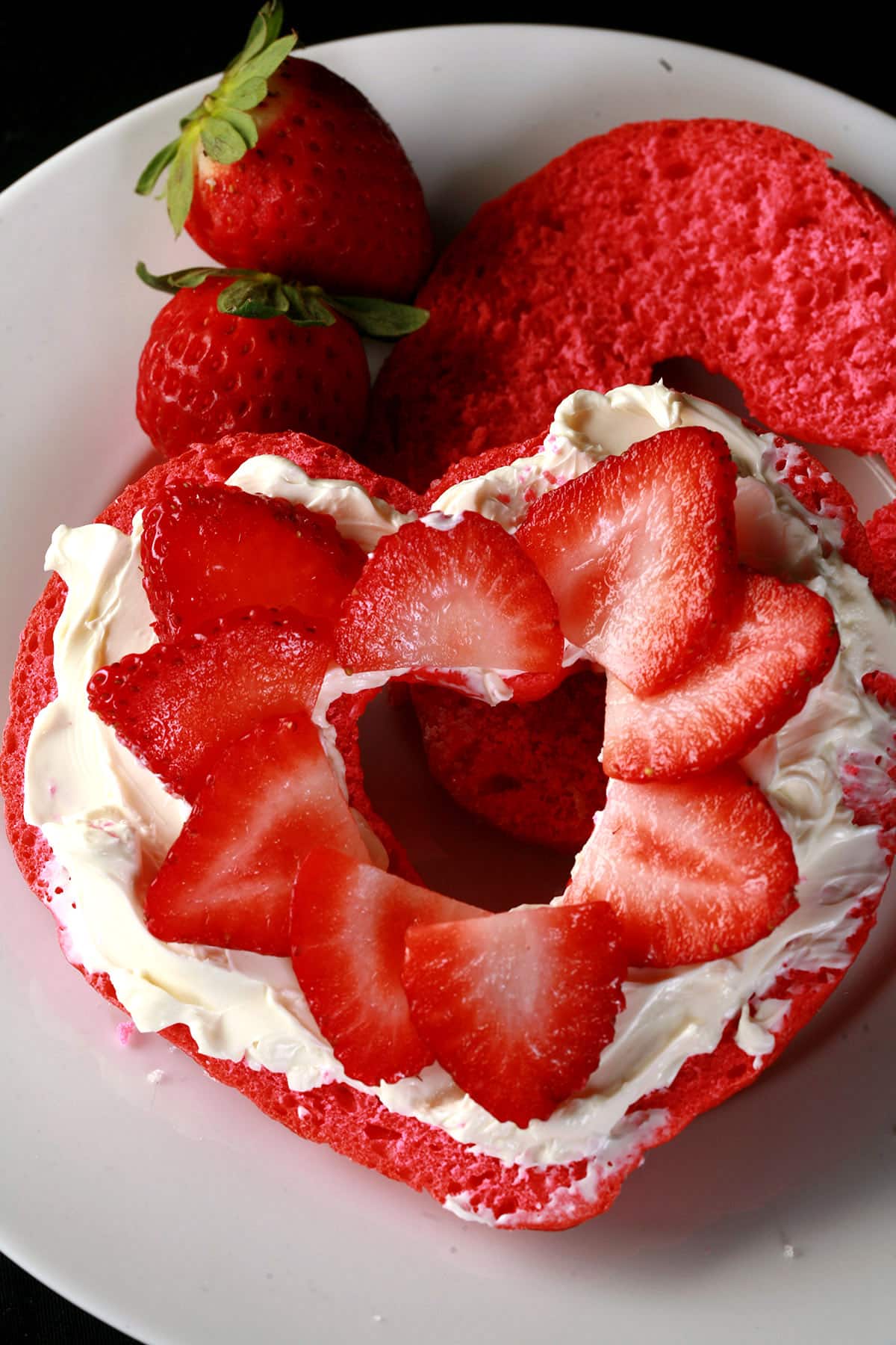 A sliced pink heart shaped bagel with cream cheese and sliced strawberries on it.