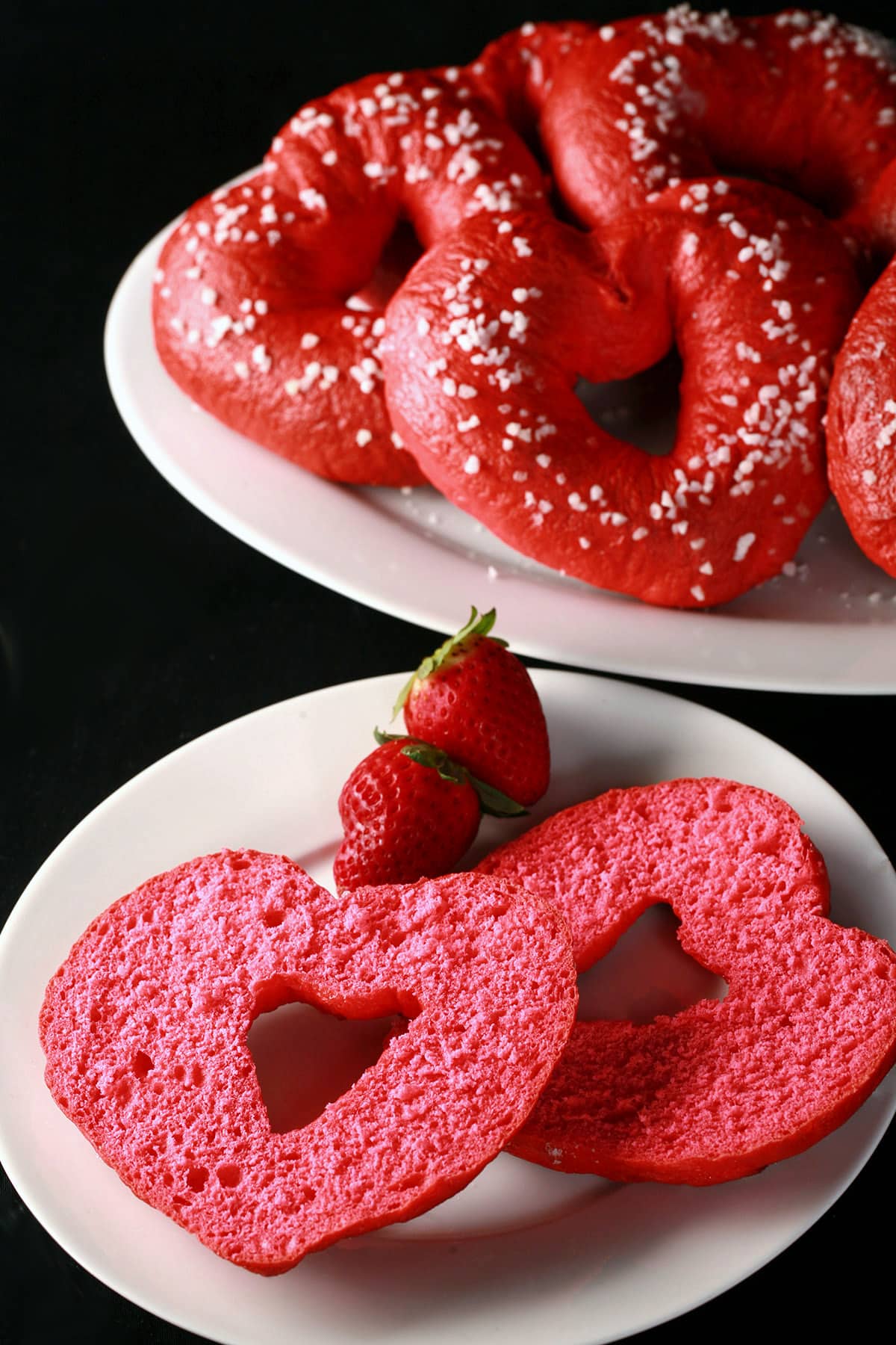 A sliced open pink heart shaped bagel on a plate, with a serving platter of valentine’s day bagels behind it.