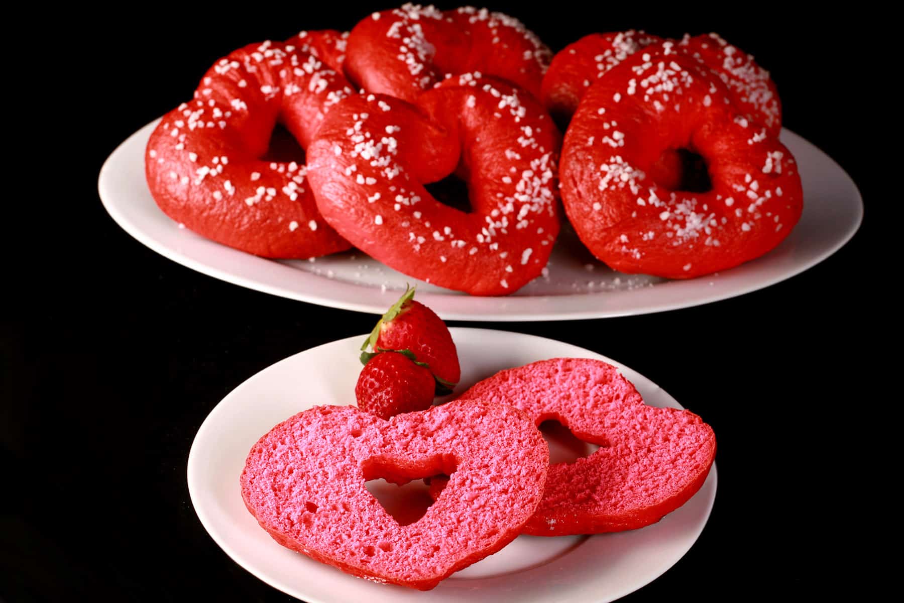 A sliced open pink heart shaped bagel on a plate, with a serving platter of valentine’s day bagels behind it.