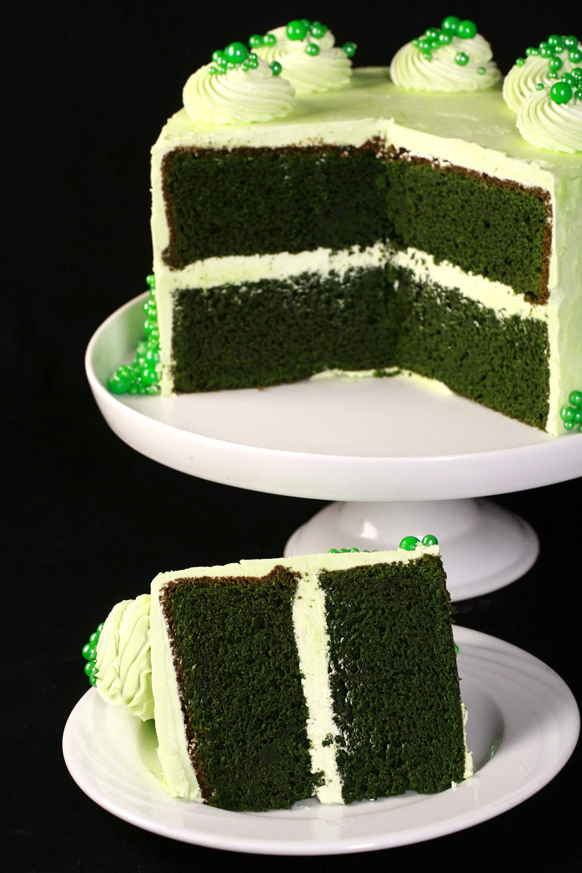 A slice of green velvet cake on a plate in front of the rest of the cake.