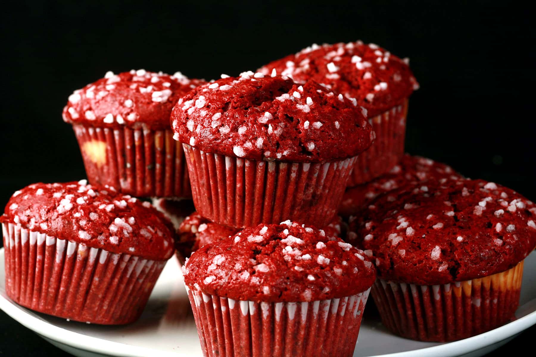 A pile of red velvet muffins on a plate, each topped with pearl sugar.