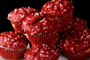 A pile of red velvet muffins on a plate, each topped with coarse sugar.