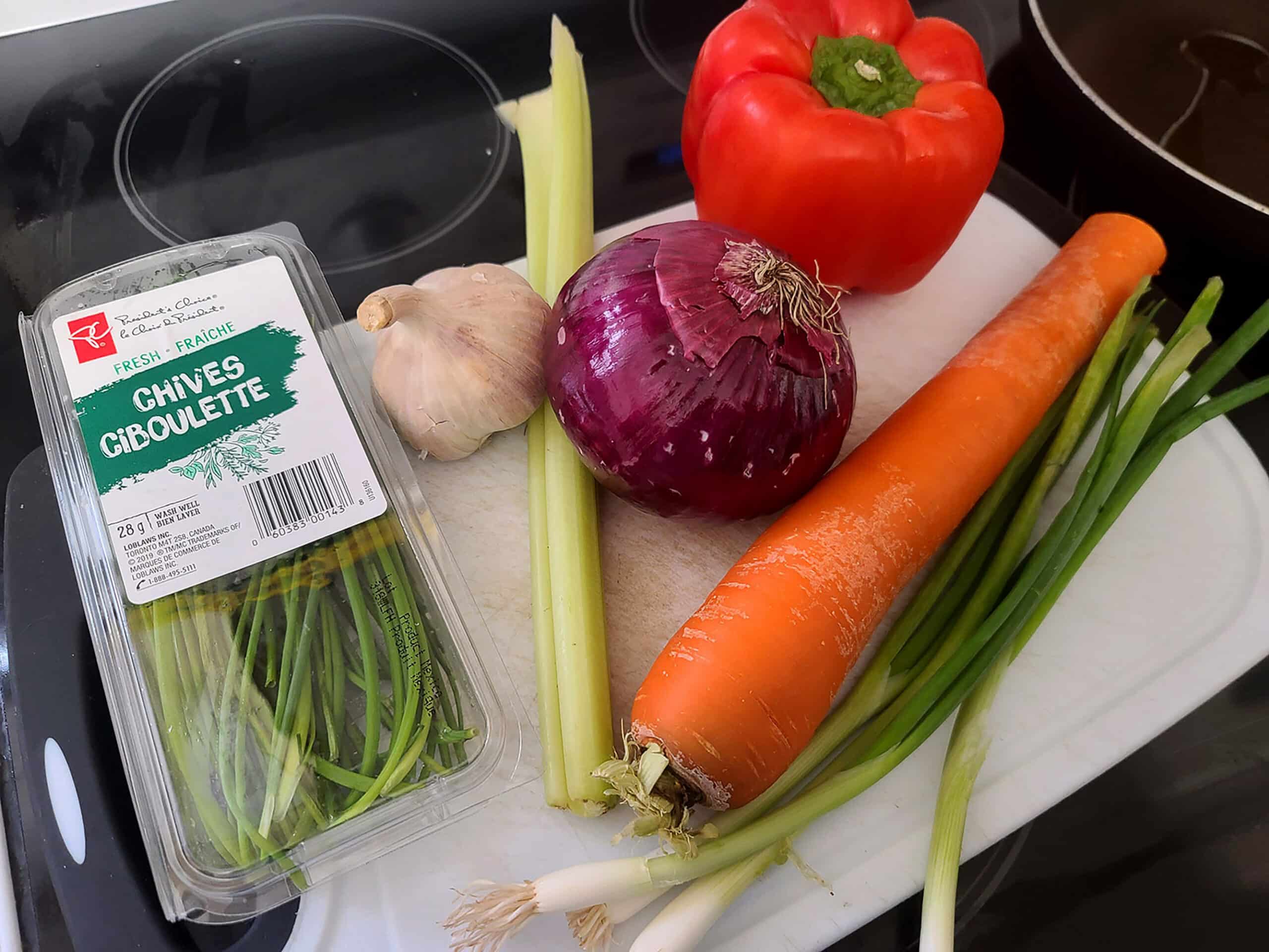 The fresh vegetables and herbs laid out on a cutting board - green onions, chives, carrot, a red onion, a garlic bulb, and a red pepper.