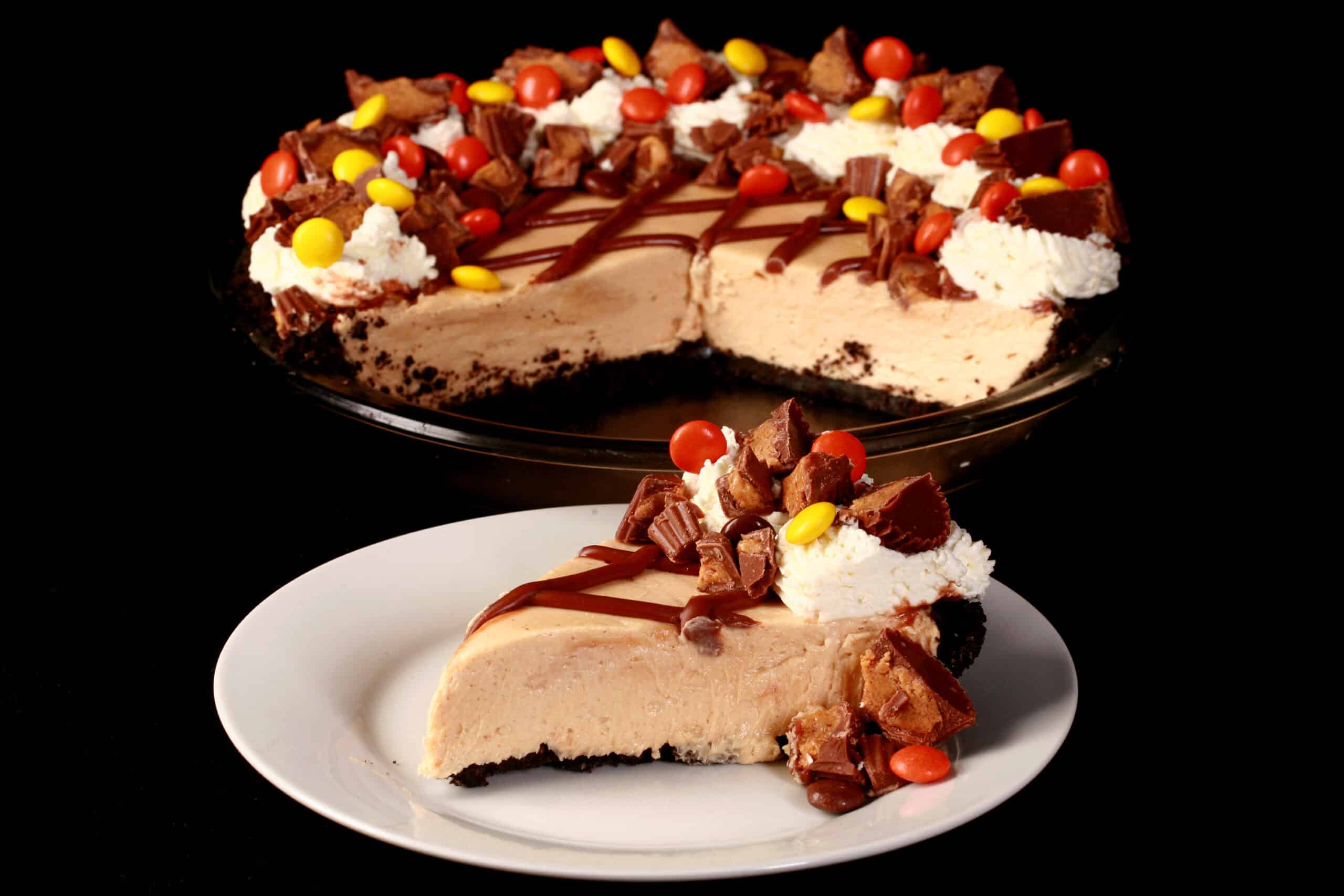 A slice of Reese’s Pie, topped with chopped up mini peanut butter cups and Reese’s Pieces. It’s sitting in front of the rest of the pie.
