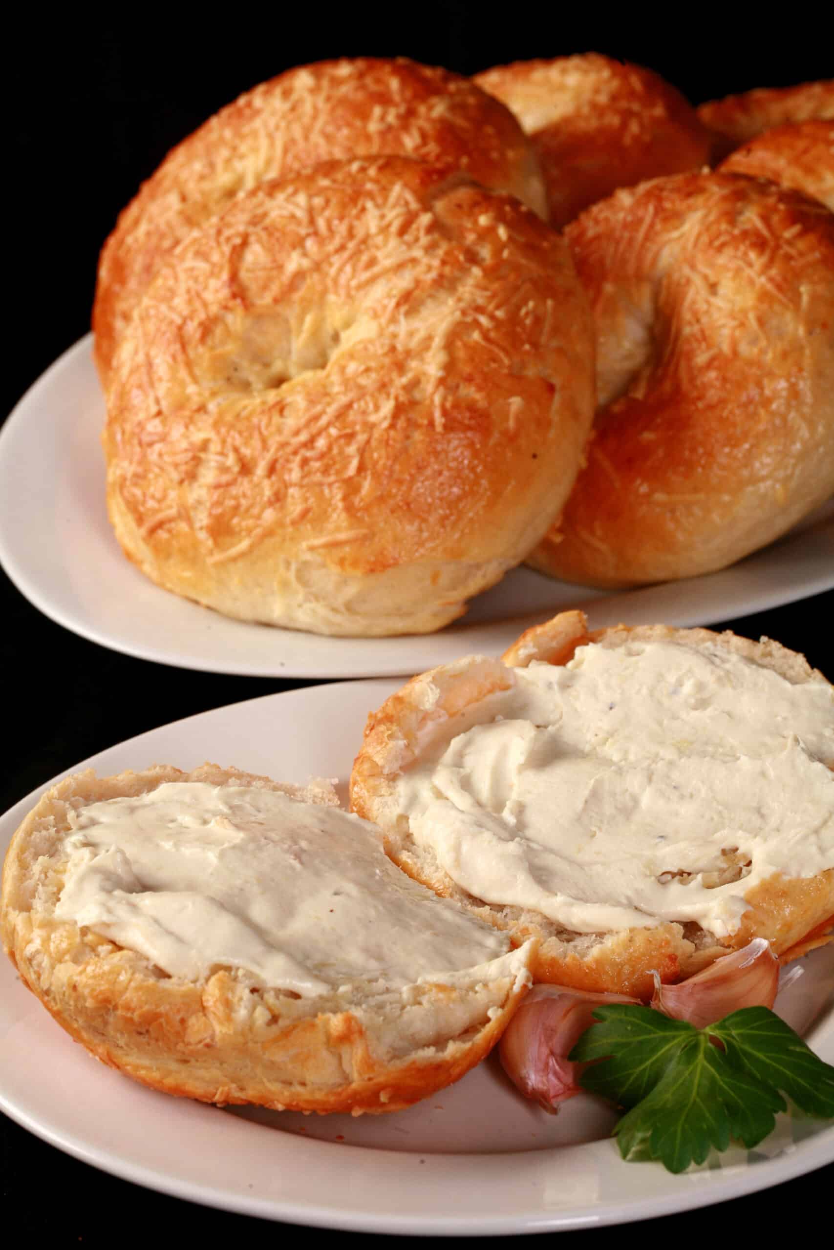 A split open roasted garlic asiago cheese bagel with cream cheese on it in front of a plate of asiago bagels.