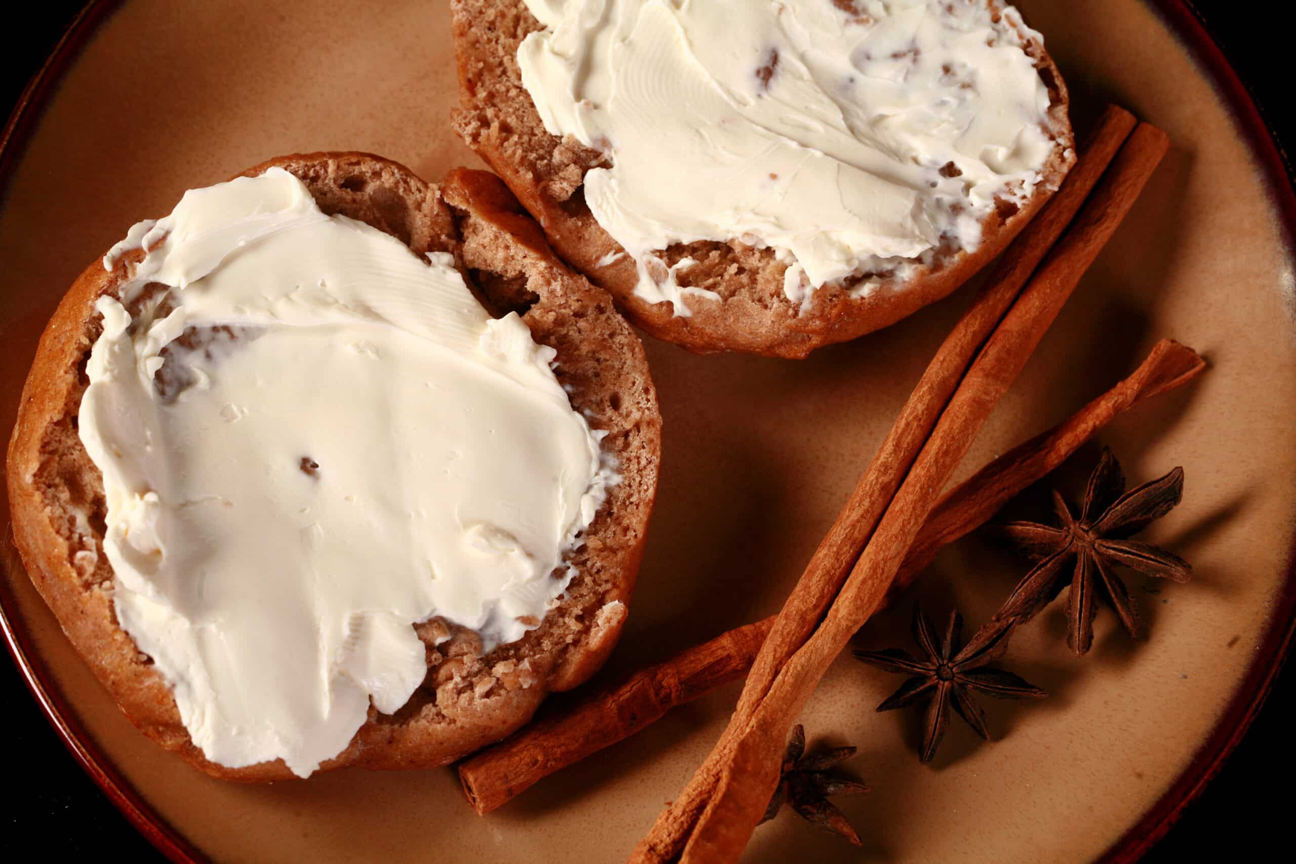 A split open chai spice bagel with cream cheese, on a plate with cinnamon sticks and star anise on the side.