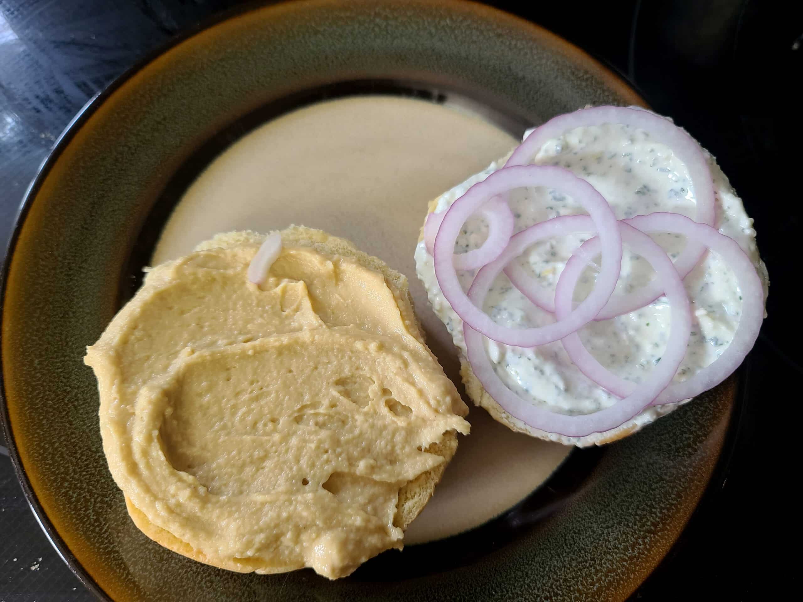 A hamburger bun prepared with hummus, red onions, and labneh feta mint spread.